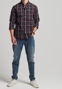 Camicia a bottoni con motivo scozzese nero e rosso, maniche arrotolate, abbinata a jeans azzurri e sneakers bianche. Design semplice con dettaglio del logo.