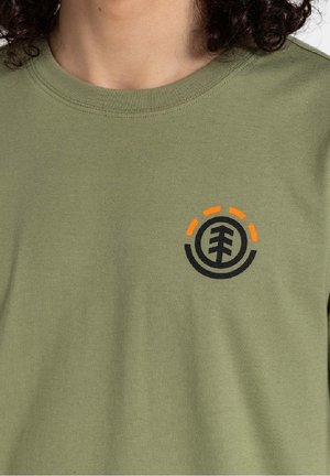 Personne portant un t-shirt vert olive à col rond avec un logo circulaire d'arbre noir et orange sur le côté gauche de la poitrine, cheveux bouclés foncés visibles au niveau du cou.