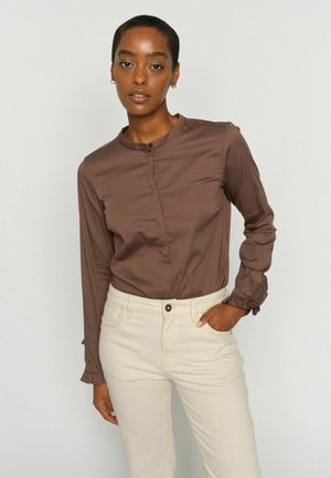 Ženska s kratkimi lasmi, oblečena v rjavo dolgoročno bluzo z nabrano manšetami in bež visoko pasnimi hlačami, stoji pred enostavno ozadje.