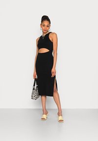 Vestido de malha preto com recorte na cintura, fendas laterais e design sem mangas. Combinado com mules amarelas de ponta aberta e uma bolsa com padrão de zebra.