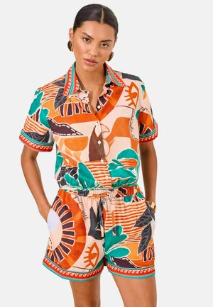 Kvinde med mørkt hår iført matchende kortærmet skjorte med krave og shorts med abstrakte orange, grønne, sorte og beige mønstre. Hænderne i lommerne.