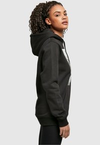 Sudadera con capucha negra, con un bolsillo frontal, puños acanalados y un diseño gráfico prominente en el pecho. Tejido suave y texturizado.