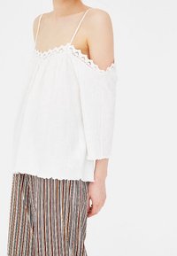 White off-shoulder top with scalloped lace trim, featuring thin straps and a relaxed fit. Paired with striped fabric pants in multiple colors.