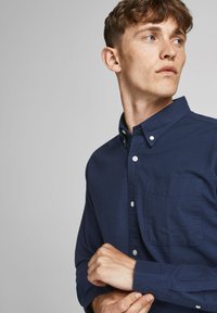 Camisa de botones azul marino hecha de algodón, con un bolsillo en el pecho, mangas largas y un cuello con detalles de botones. Textura suave.