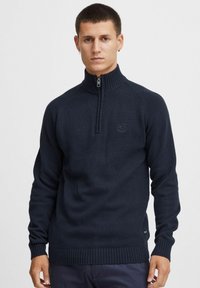 Sweater zippé bleu marine avec un col haut, des poignets et un ourlet côtelés, texture tricotée et logo brodé sur la poitrine.