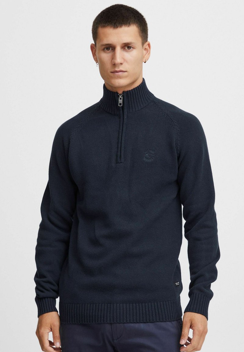 Sweater zippé bleu marine avec un col haut, des poignets et un ourlet côtelés, texture tricotée et logo brodé sur la poitrine.