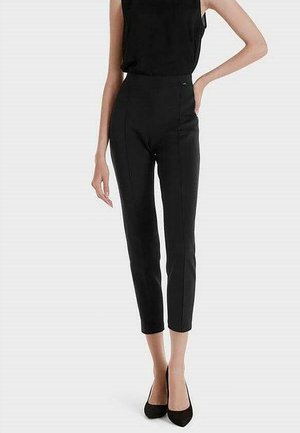 Femme portant un top noir ajusté sans manches, un pantalon slim noir taille haute coupé au-dessus des chevilles, et des escarpins noirs à bout pointu.
