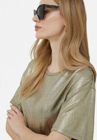 Camicia corta in oro metallico con texture a coste, indossata con occhiali da sole neri con una montatura decorata. I capelli sono lunghi e lisci.