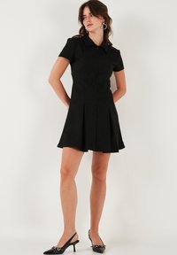 Čierne šaty s krátkym rukávom, ktoré majú golier, skladanú sukňu a hladký povrch. Kombinované s čiernymi slingback topánkami na podpätku a kruhovými náušnicami.