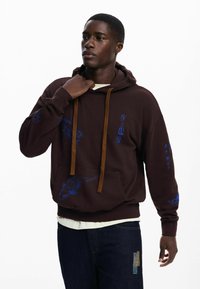 Brauner Pullover-Hoodie mit blauer Stickerei, darunter florale und fischartige Muster. Braune Kordeln und eine Fronttasche. Dunkle Jeans darunter.