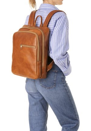 Sac à dos en cuir brun avec deux compartiments, poche zippée à l'avant et bretelles, présenté sur une chemise rayée et un jean.
