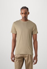 Beige t-shirt med korte ærmer lavet af glat stof, med en rund halsudskæring og et lille logo på brystet; iført med lyse beige bukser.