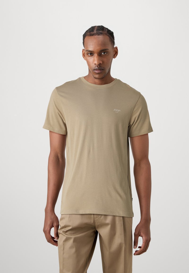 Beige t-shirt med korte ærmer lavet af glat stof, med en rund halsudskæring og et lille logo på brystet; iført med lyse beige bukser.