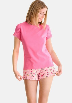 Femme portant un haut rose à manches courtes avec bordure en dentelle et un short rose assorti à motif floral, ajustant l'ourlet du short.