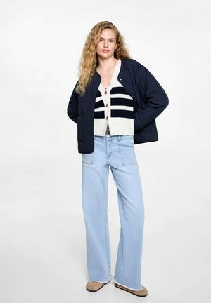 Giacca trapuntata blu navy sopra un cardigan bianco con righe blu navy, jeans a gamba larga blu e scarpe color sabbia. L'outfit presenta bottoni e orli sfrangiati.