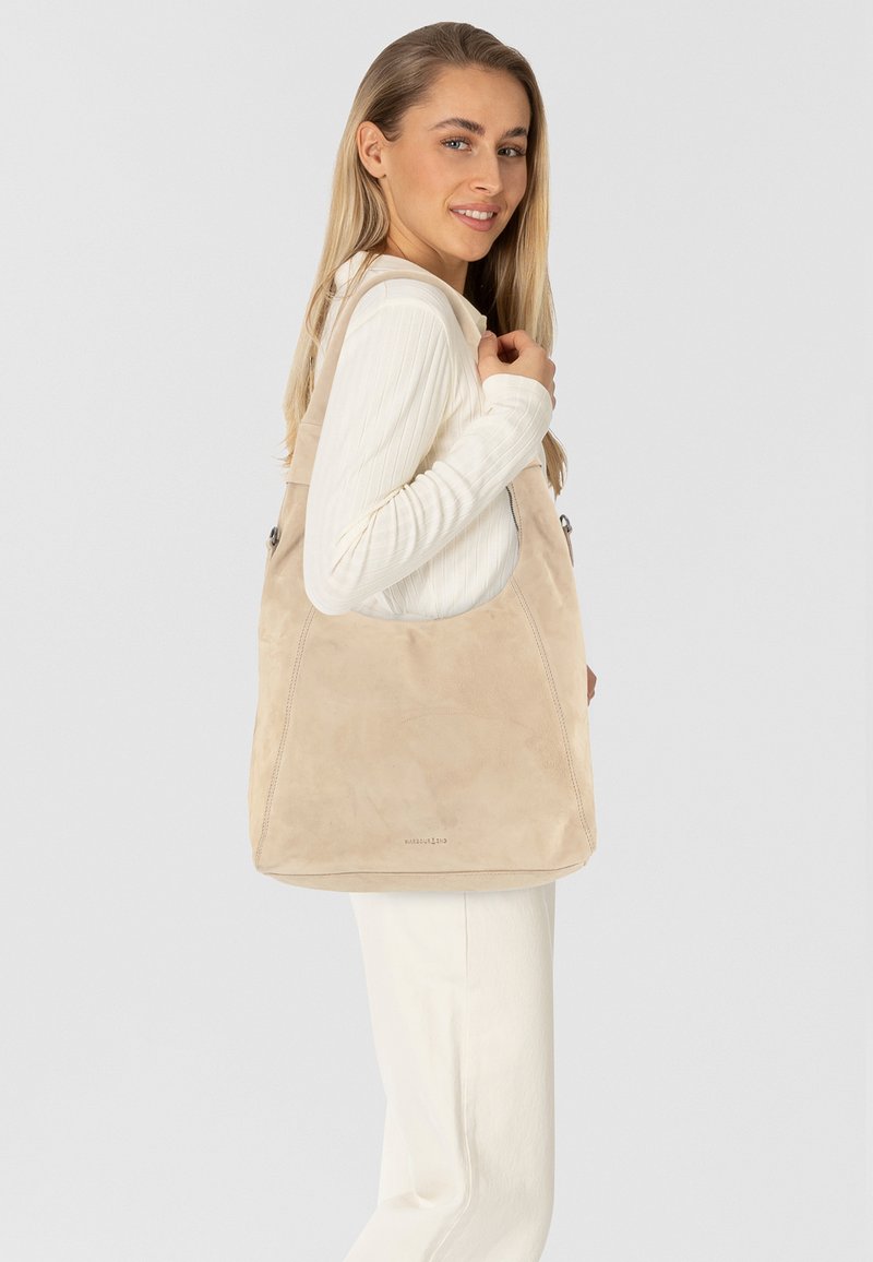 Sac à épaule en daim beige avec une texture lisse, ouverture sur le dessus et des détails cousus. Le sac possède un design large et semi-circulaire ainsi que des accents en métal.