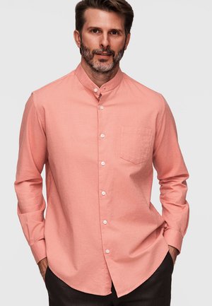 Mężczyzna z brodą, ubrany w koralowo-różową koszulę na długi rękaw z kołnierzykiem mandarynkowym, ręce częściowo w kieszeniach ciemnych spodni.