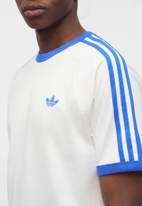 Person wearing a white Adidas shirt with blue trim, three blue stripes on the shoulder, and a blue Adidas logo on the chest.