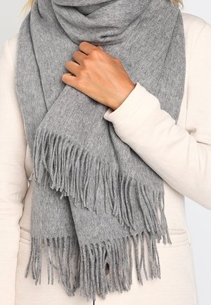 Personne portant un manteau beige tenant une grande écharpe grise douce avec de longs franges enroulée autour du cou.