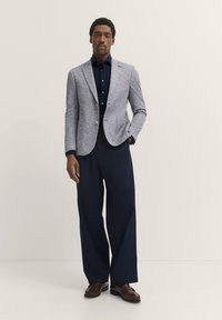 Hellgrauer, strukturierter Blazer über einem marineblauen Hemd, kombiniert mit weit geschnittenen, marineblauen Hosen und braunen Loafers. Schlichte Knöpfe betonen das Hemd.