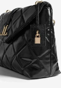 Musta tikattu nahkainen käsilaukku, jossa on kultainen padlock-yksityiskohta ja logovarustus läpällä, näyttäen sivukulman näkymän.