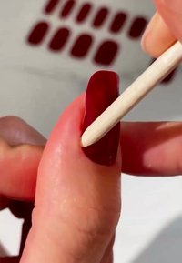 Smalto rosso applicato a un'unghia, con un bastoncino di legno utilizzato per i dettagli. Sullo sfondo si vedono ulteriori pezzi di unghie rosse.