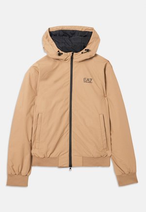 Beige lett jakke med hette, glidelås, elastiske mansjetter og sidelommer; har en svart innerforing og logo på brystet.