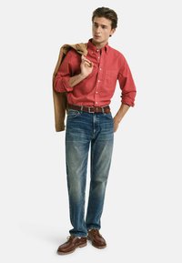 Rotes Hemd mit Knöpfen, blaue verwaschene Jeans, braune Lederschuhe und eine sandfarbene Jacke über die Schulter gelegt. Legeres Ensemble mit kontrastierenden Farben.