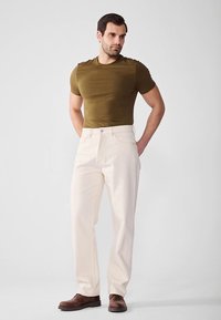 Olivengrøn tætsiddende t-shirt, hvide bukser med lige ben, brune sko. T-shirten har tekstur; bukserne har klassiske lommer og lynlås.