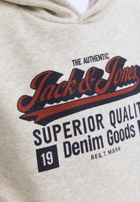 Bunda s kapuco v svetlo bež barvi s temno modrim in rjavim napisom "Pravi Jack & Jones Superior Quality Denim Goods 19".