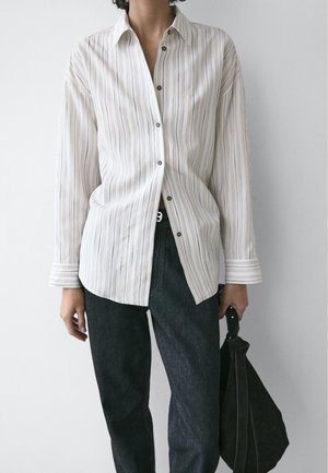 Personne portant une chemise blanche à rayures verticales et un pantalon noir, tenant un sac à main noir devant un fond uni.