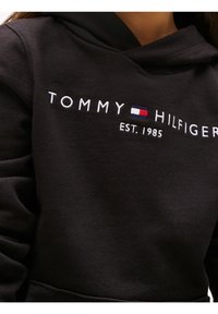 Sweat à capuche noir avec texte blanc "Tommy Hilfiger", logo rouge, blanc et bleu, et "EST. 1985" brodé sur la poitrine.