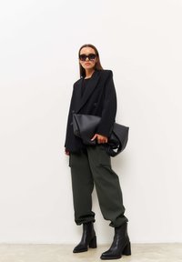Blazer noir oversize à double boutonnage, haut noir ajusté, pantalon cargo vert olive et bottines noires à talons épais. Les accessoires incluent de grandes lunettes de soleil.