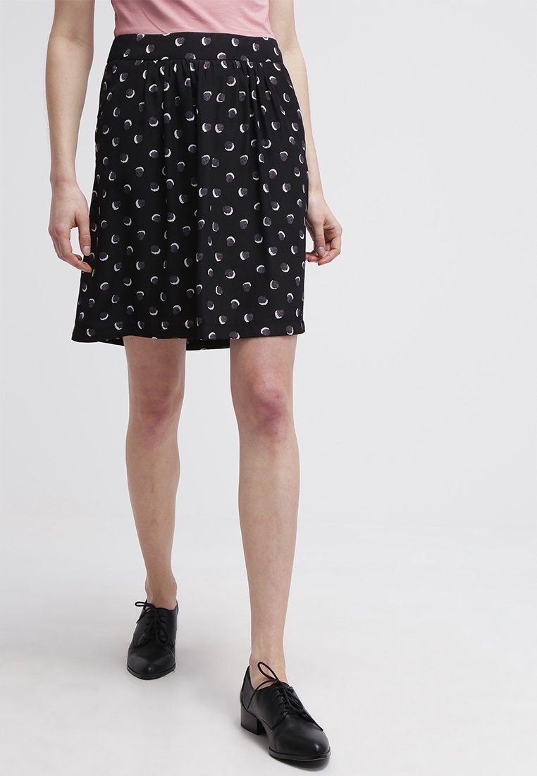 Femme portant une jupe noire avec des motifs circulaires blancs, un haut rose et des chaussures noires à lacets, marchant devant un fond uni clair.