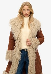 Brun falsk suede frakke med en lang, fluffy beige pelskrave og manchetter, som har et båndlukning og en struktureret beige sweater nedenunder.