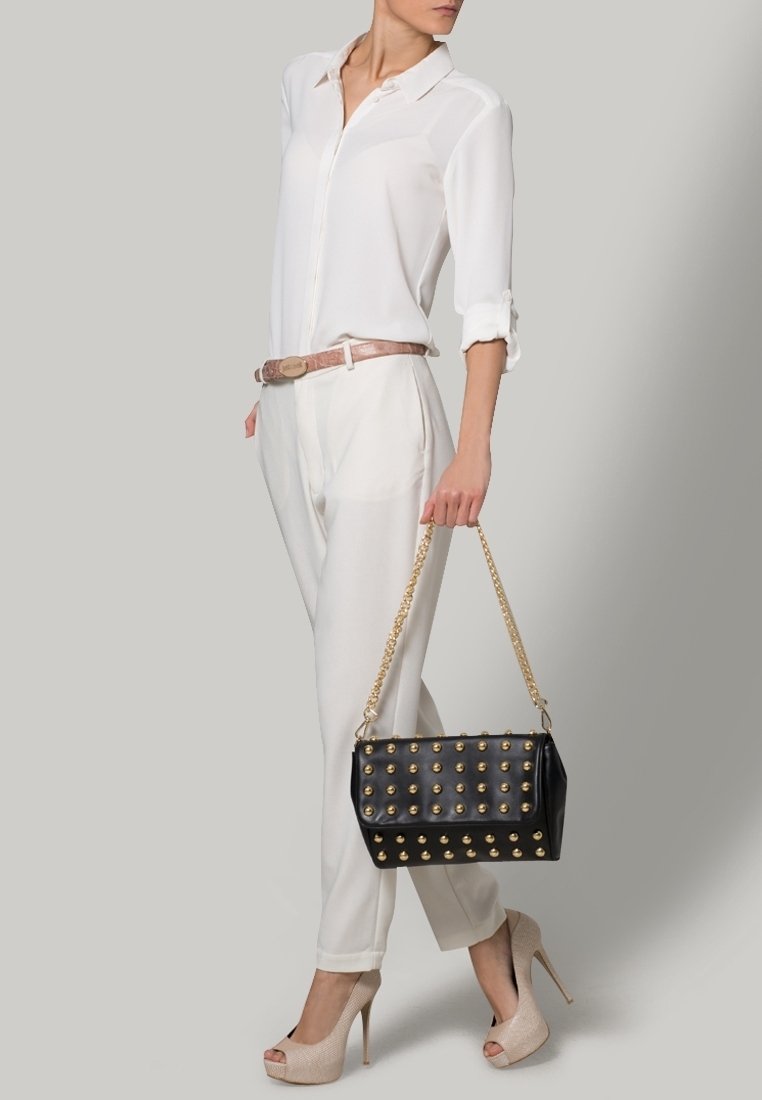 Femme en blouse blanche et pantalon, talons beige, tenant un sac à main noir avec clous dorés et bandoulière chaîne, debout devant un fond uni.