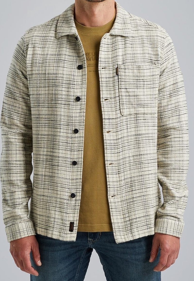 Homme portant une chemise à carreaux crème et noire avec des boutons, par-dessus un t-shirt moutarde et un jean bleu, debout les mains détendues sur les côtés.