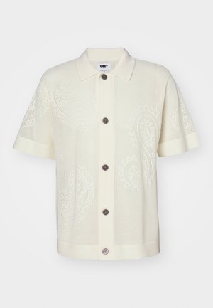 Cream short-sleeve collared shirt with a patterned knit design, featuring five brown buttons and a pink accent button on the lower placket.