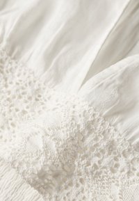 Weißes Kleidungsstück mit strukturiertem Stoff und aufwändigen Spitzenverzierungen in der Mitte. Es hat weiches, geplättetes Material, das die zarte Handwerkskunst hervorhebt.