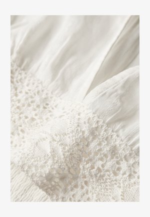 Hvidt tøj med struktureret stof og intrikat blonderdetalje i midten. Har blødt, plisseret materiale, som fremhæver delikat håndværk.