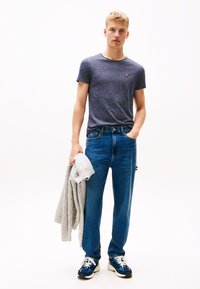 Mladý muž v modrém tričku a džínách, držící v jedné ruce šedý pletený svetr, stojící proti jednoduchému bílému pozadí.