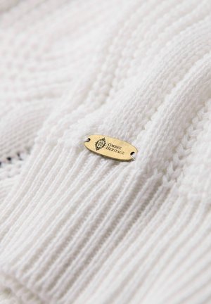 Tissu tricoté blanc avec une texture côtelée, doté d'une étiquette en or gravée avec "OMBRE HERITAGE" affichée de manière proéminente.