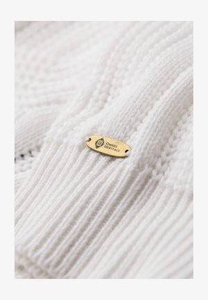 Weißer gestrickter Stoff mit einer gerippten Textur, auf dem ein goldener Anhänger mit der Gravur "OMBRE HERITAGE" deutlich sichtbar ist.