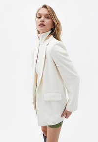 Blazer in tessuto bianco con una silhouette aderente, tasca frontale singola e spalle strutturate, abbinato a un maglione a collo alto e pantaloni shorts.