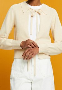 Personne portant un cardigan beige avec fermeture nœud papillon sur un haut blanc et un pantalon blanc taille haute, mains jointes, sur fond orange.