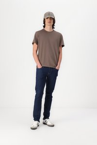 Tricou din bumbac maro, blugi din denim, șapcă gri și adidași albi cu dungi negre. Modelul stă pe un fundal alb simplu.