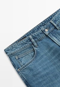 Blå denimjeans, der viser taljebåndet, forlommen, bæltestropperne og metal knaplukningen på en hvid baggrund.