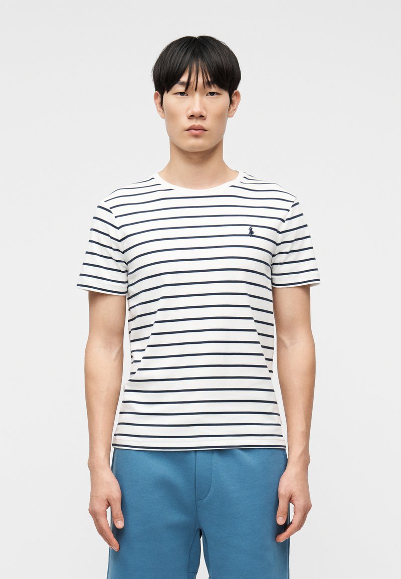 Νέος άνδρας που φοράει λευκό και ναυτικό μπλε ριγέ μπλουζάκι και μπλε παντελόνι, στέκεται μπροστά σε απλό φωτεινό φόντο, κοιτώντας προς τα εμπρός.