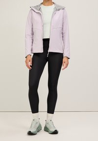 Ljust lila softshell-jacka med grå huva, framtill dragkedja och två sidofickor, tillsammans med svarta leggings och ljusgröna skor.