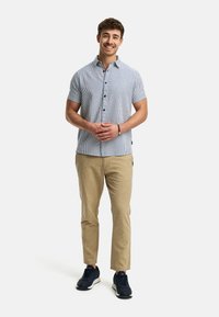 Kortærmet skjorte med knap i blå og hvide striber, beige bukser og mørke sneakers, står med hænderne flettet, neutral baggrund.